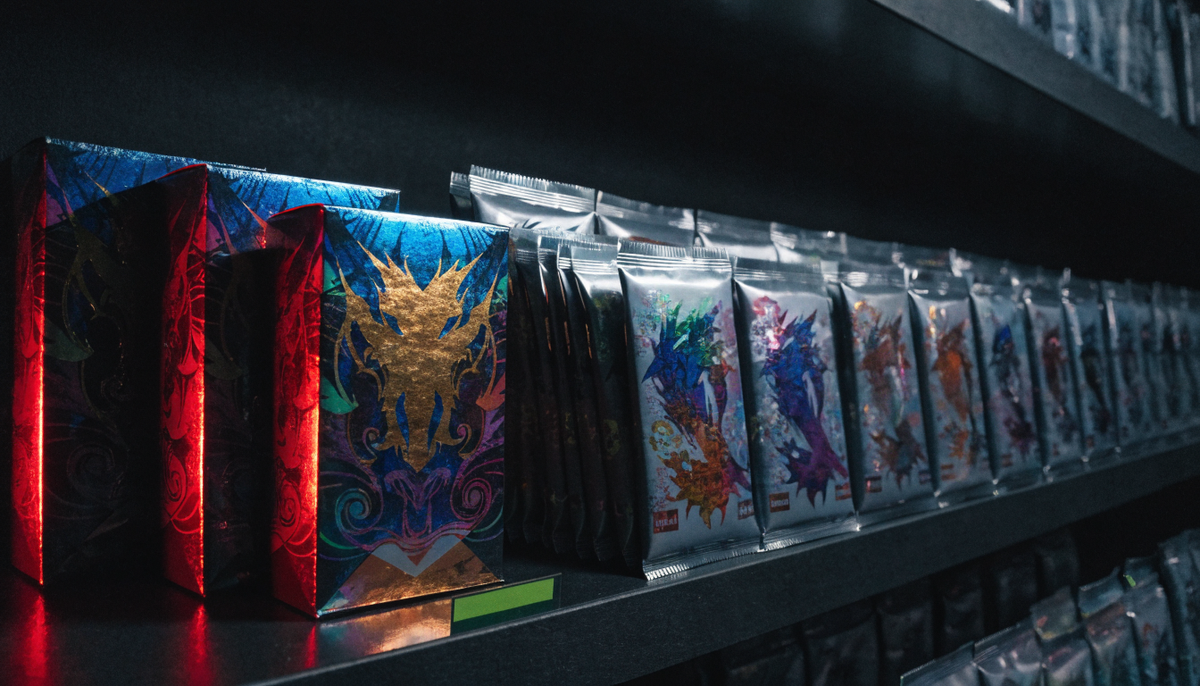 Sealed trading card booster boxes and packs stacked on a dark shop shelf with one empty gap where stock has sold out